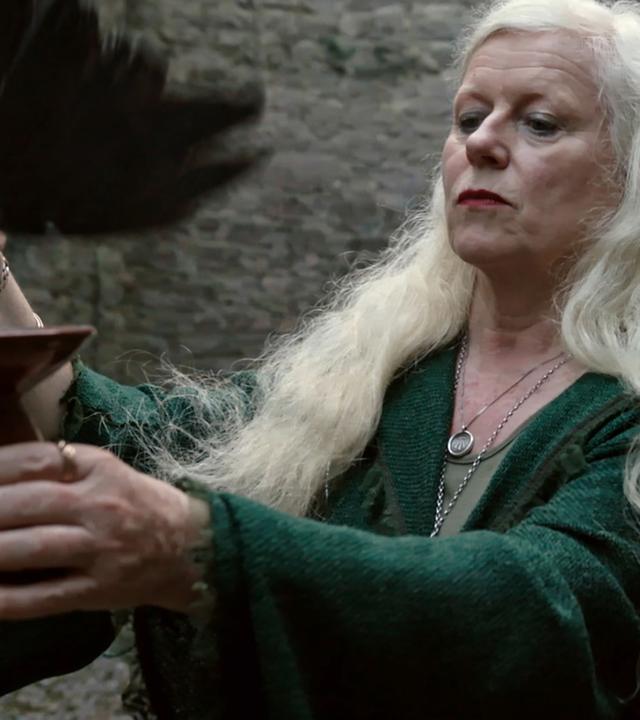 Eimear Burke mit langen, grau-blonden Haaren hält einen Kelch in der einen Hand und eine schwarze Feder in der anderen.