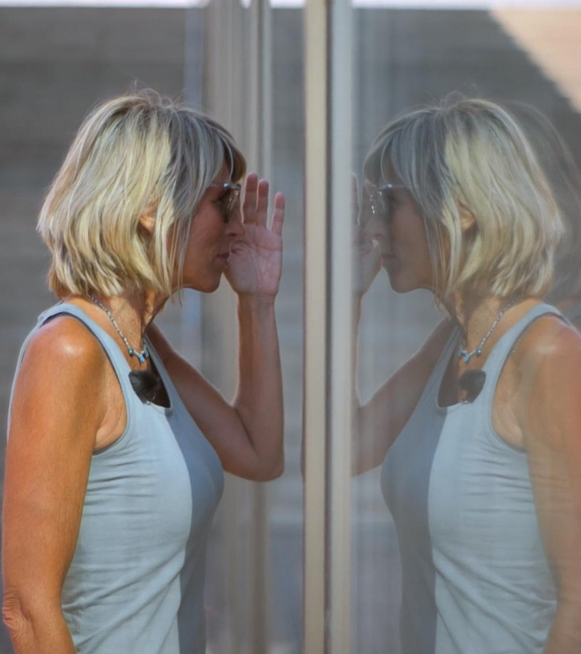 Steffi, eine sportlich schlanke Frau mit schulterlangen blonden Haaren steht an einer Glasscheibe und schaut hinein. Sie trägt eine Sonnenbrille und ein blaues Tank-Top. Neben ihr ist ihr Spiegelbild zu sehen.