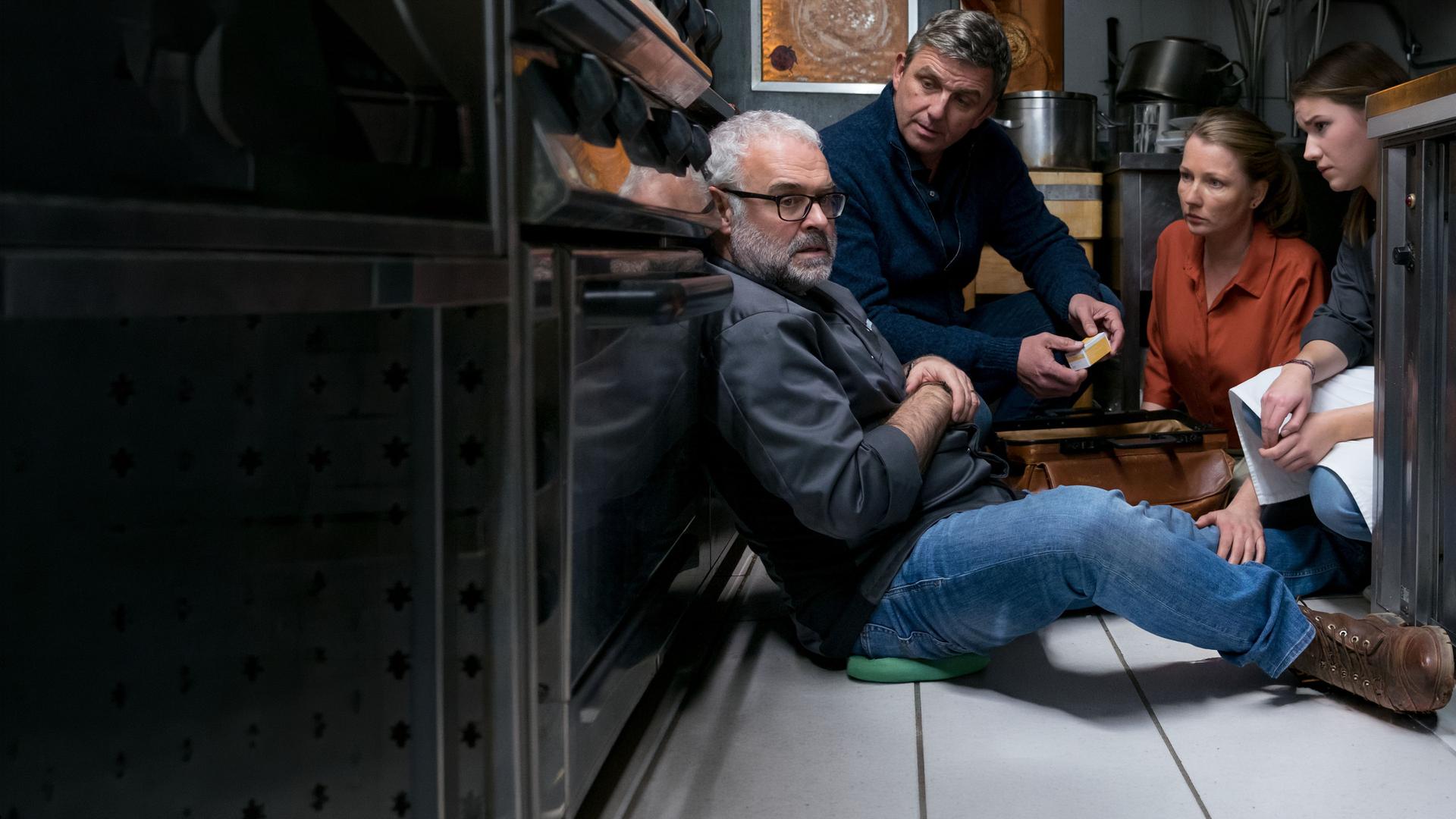 Thomas Bühler (Jophi Ries) sitzt in einer Großküche auf dem Boden und blickt zur Seite. Vor ihm knien Dr. Martin Gruber (Hans Sigl), Frau Tine (Katja Studt) und Kara (Carolin Garnier) und sehen ihn besorgt an. 