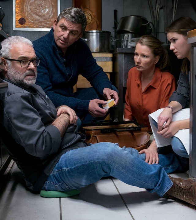 Thomas Bühler (Jophi Ries) sitzt in einer Großküche auf dem Boden und blickt zur Seite. Vor ihm knien Dr. Martin Gruber (Hans Sigl), Frau Tine (Katja Studt) und Kara (Carolin Garnier) und sehen ihn besorgt an. 