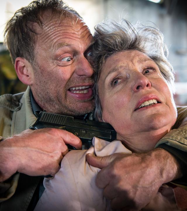 "Notruf Hafenkante - Die falsche Frau": Porträt von Andreas Beckmann (Rainer Strecker), der Marlies Hansen (Peggy Lukac) mit dem linken Arm im Schwitzkasten hält, mit der rechten Hand eine Waffe an den Kopf hält.