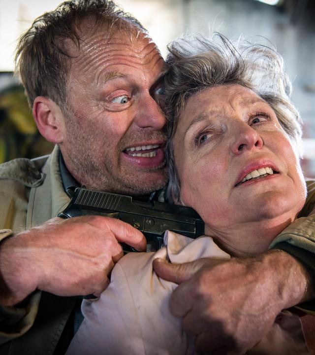 "Notruf Hafenkante - Die falsche Frau": Porträt von Andreas Beckmann (Rainer Strecker), der Marlies Hansen (Peggy Lukac) mit dem linken Arm im Schwitzkasten hält, mit der rechten Hand eine Waffe an den Kopf hält.