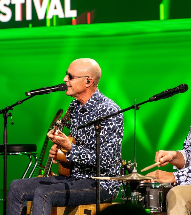 Die feisten sitzen auf Barhockern auf der Bühne. Vor ihnen stehen Mikrofonständer. Der Hintergrund ist grün beleuch