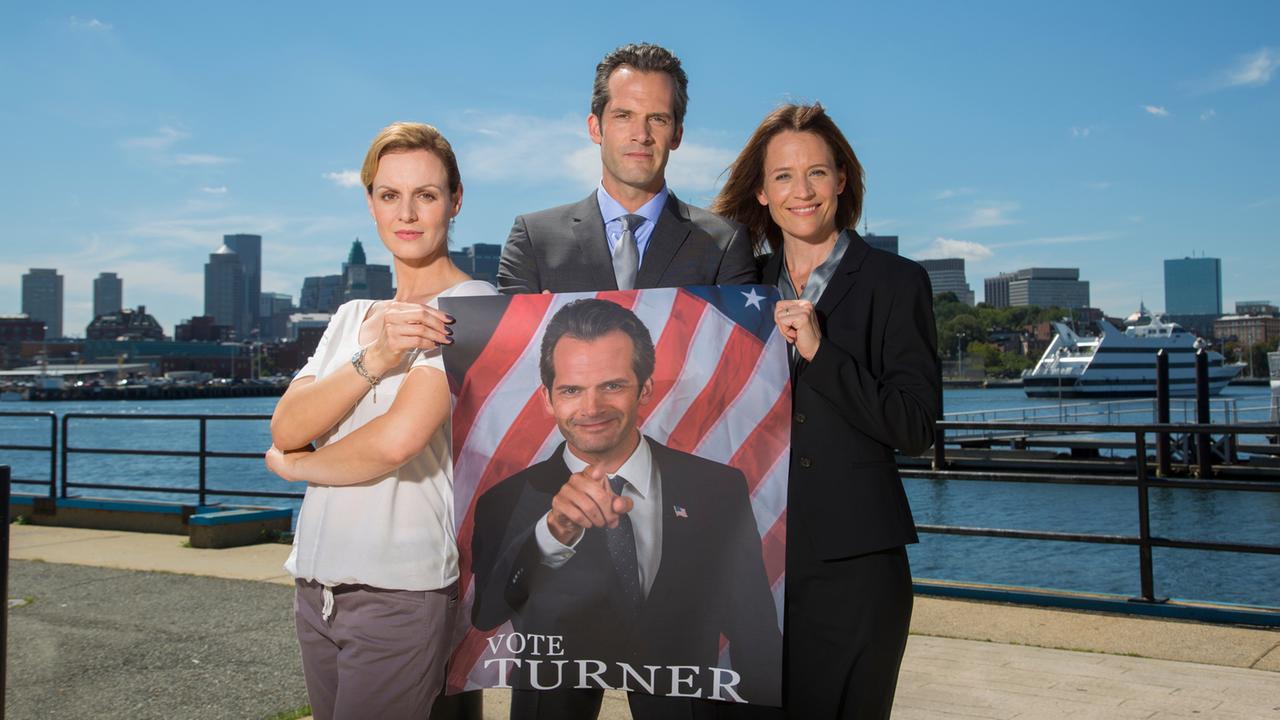 Pressefrau Robin Jennings (Anja Nejarri), Eric Turner (Martin Rapold) und seine Frau Julie (Regula Grauwiller) stehen vor der Bostoner Skyline und halten sein Wahlplakat in den Händen.