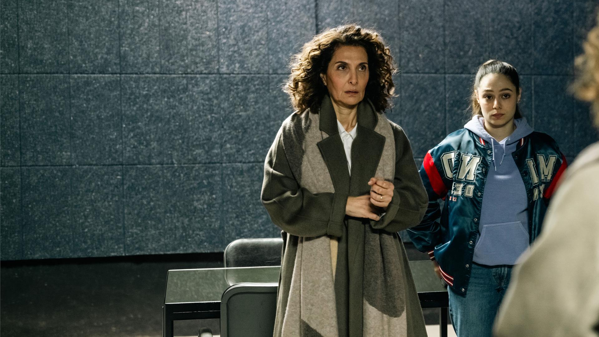 "SOKO Köln - Die Frau auf dem roten Fahrrad": Yasmin Barani (Proschat Madani) steht neben Amira Mahdi (Aylin Ravanyar) vor einer dunkelgrauen Wand mit Fliesenmuster. Sie blickt angespannt nach oben. Amira hat die Arme in die Hüften gestemmt und sieht ernst zu Lilly Funke (Tatjana Kästel), die ihr gegenüber steht, mit dem Rücken zur Kamera.