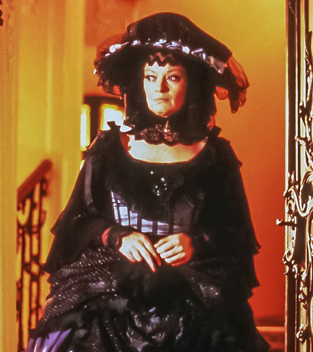 Die skeptische Sorgenfee (Jana Brejchová) steht im Rahmen einer geöffneten, reich verzierten Tür. Beide Türflügel sind geöffnet, hinter ihr erstrahlt warmes Licht. Sie tritt ins Dunkle hinaus.