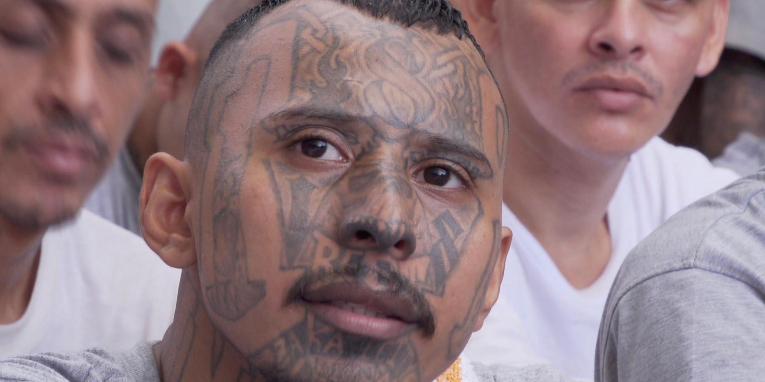 Ein junger Mann mit tätowiertem Gesicht und grauem T-Shirt sitzt neben anderen Männern mit Tattoos.