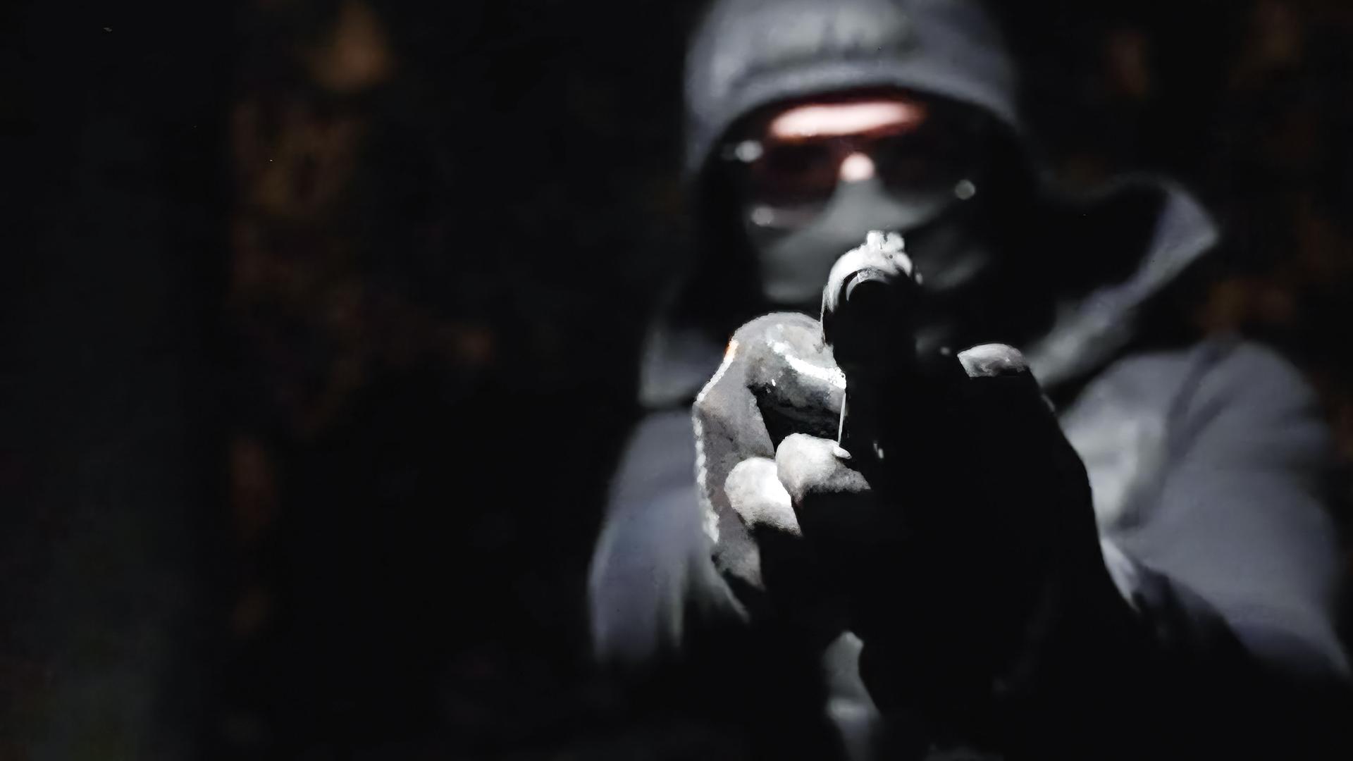 In der Dunkelheit hält ein vermummter Mann eine Waffe in den Händen und zielt damit geradeaus.