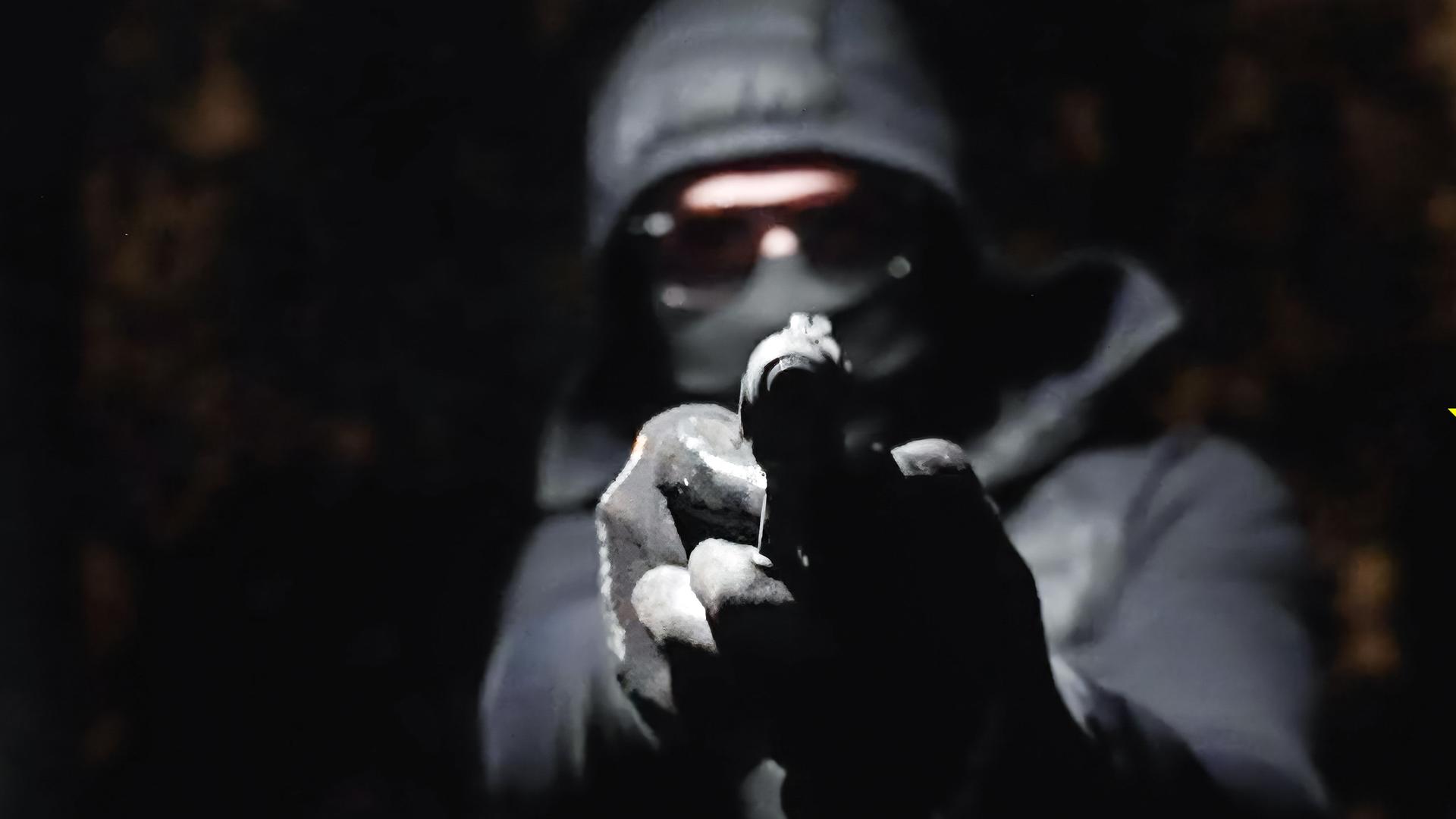 In der Dunkelheit hält ein vermummter Mann eine Waffe in den Händen und zielt damit geradeaus.