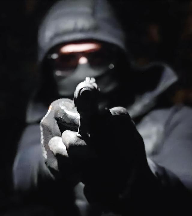 In der Dunkelheit hält ein vermummter Mann eine Waffe in den Händen und zielt damit geradeaus.