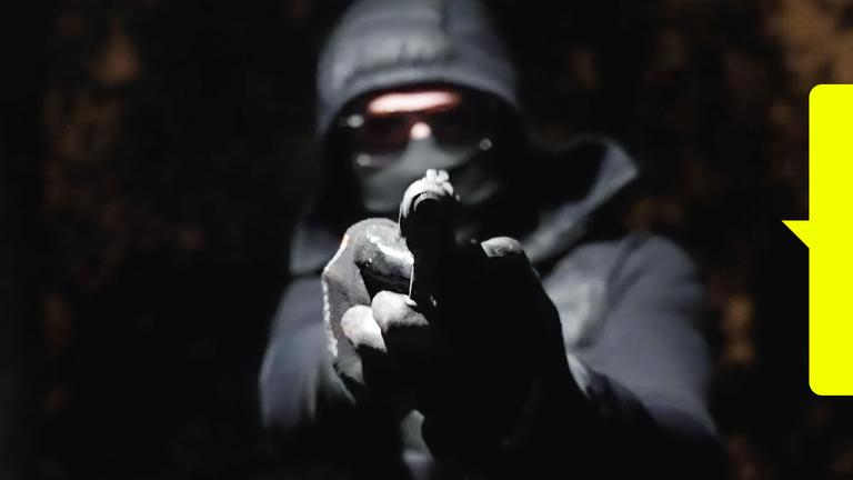 In der Dunkelheit hält ein vermummter Mann eine Waffe in den Händen und zielt damit geradeaus.