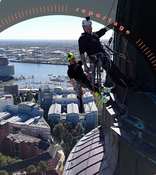 Industriekletterer Eric Bossaller und Janis Weber hängen an dem Turm des Hamburger Michels. Mit jeweils einer Hand haten sie sich an ihren Seilen fest und schauen, versetzt voneinander, lächelnd in die Kamera. Sie tragen mit Go-Pro ausgestatteten Helme und weiteres Zubehör an ihren Hüftgürteln. Teile der Elbe und der Stadt liegen im Hintergrund.