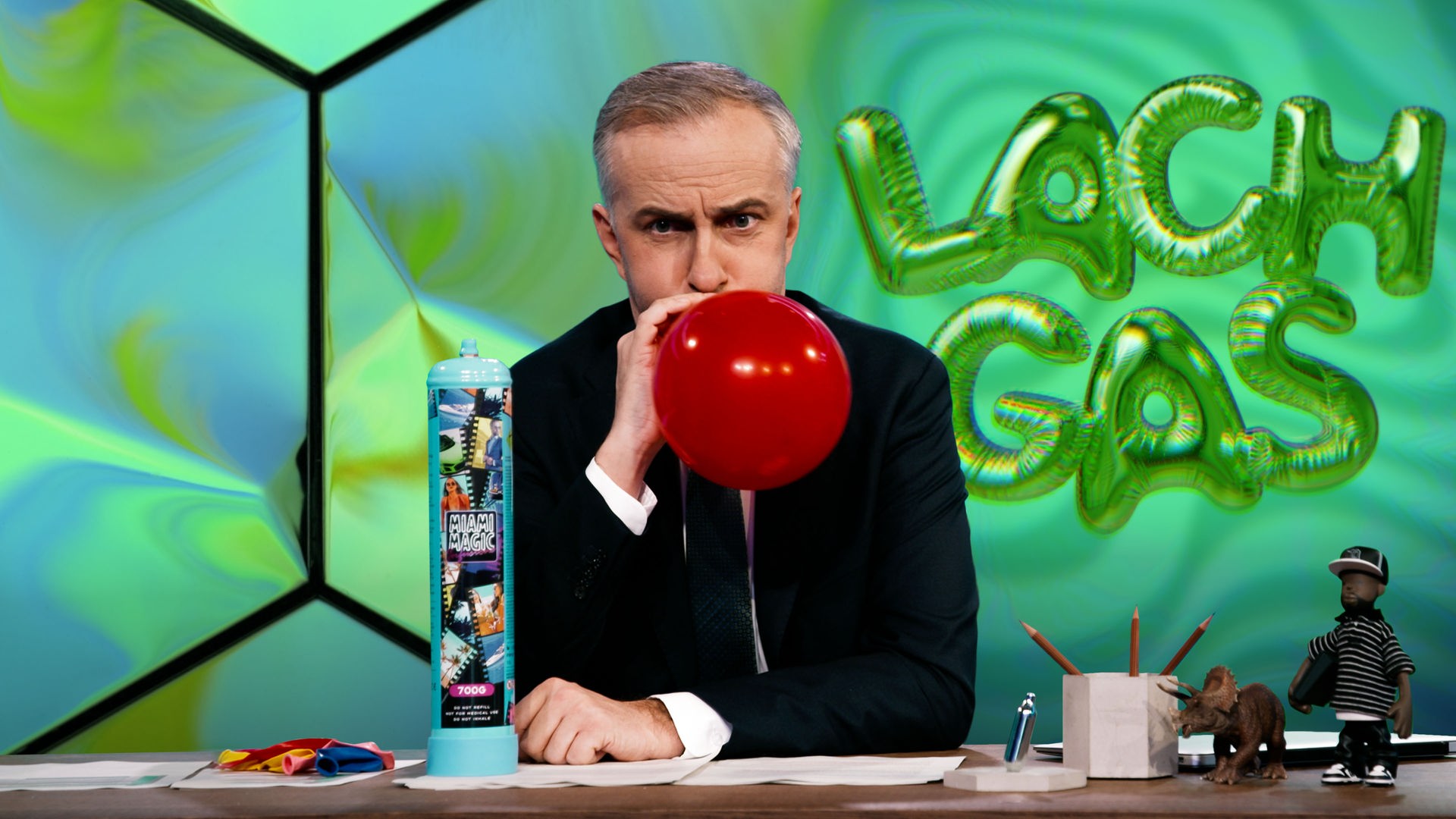 Jan Böhmermann sitzt am Schreibtisch und hält einen roten, aufgeblasenen Ballon an den Mund. Links neben ihm steht eine blaue Gasflasche mit der Aufschrift "Miami Magic". Rechts im Bild ist der Schriftzug "Lachgas" in grünen, ballonartigen Buchstaben abgebildet.