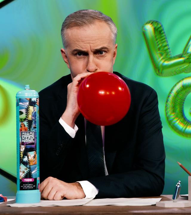 Jan Böhmermann sitzt am Schreibtisch und hält einen roten, aufgeblasenen Ballon an den Mund. Links neben ihm steht eine blaue Gasflasche mit der Aufschrift "Miami Magic". Rechts im Bild ist der Schriftzug "Lachgas" in grünen, ballonartigen Buchstaben abgebildet.