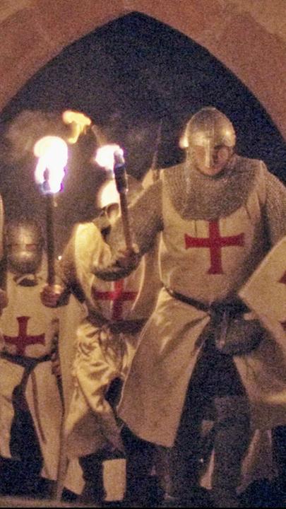 "Die Geheimnisse der Tempelritter: Aufstieg zum Wirtschaftsimperium": Nachgestellte Szene: Im Licht brennender Fackeln stürmen vier Tempelritter in Rüstung durch ein Tor frontal auf die Kamera zu.