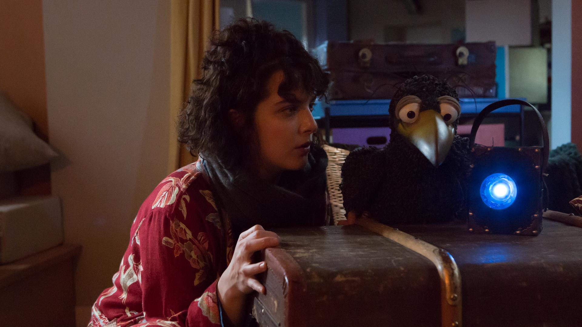 Es ist dunkel in Siebensteins Laden. Siebenstein schaut gespannt zu Rudi, der neben ihr hockt und eine blau leuchtende Lampe in den Laden richtet. Im Hintergrund liegt Koffer auf dem Tresen und schaut zu den beiden.