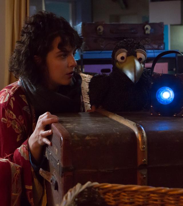 Es ist dunkel in Siebensteins Laden. Siebenstein schaut gespannt zu Rudi, der neben ihr hockt und eine blau leuchtende Lampe in den Laden richtet. Im Hintergrund liegt Koffer auf dem Tresen und schaut zu den beiden.