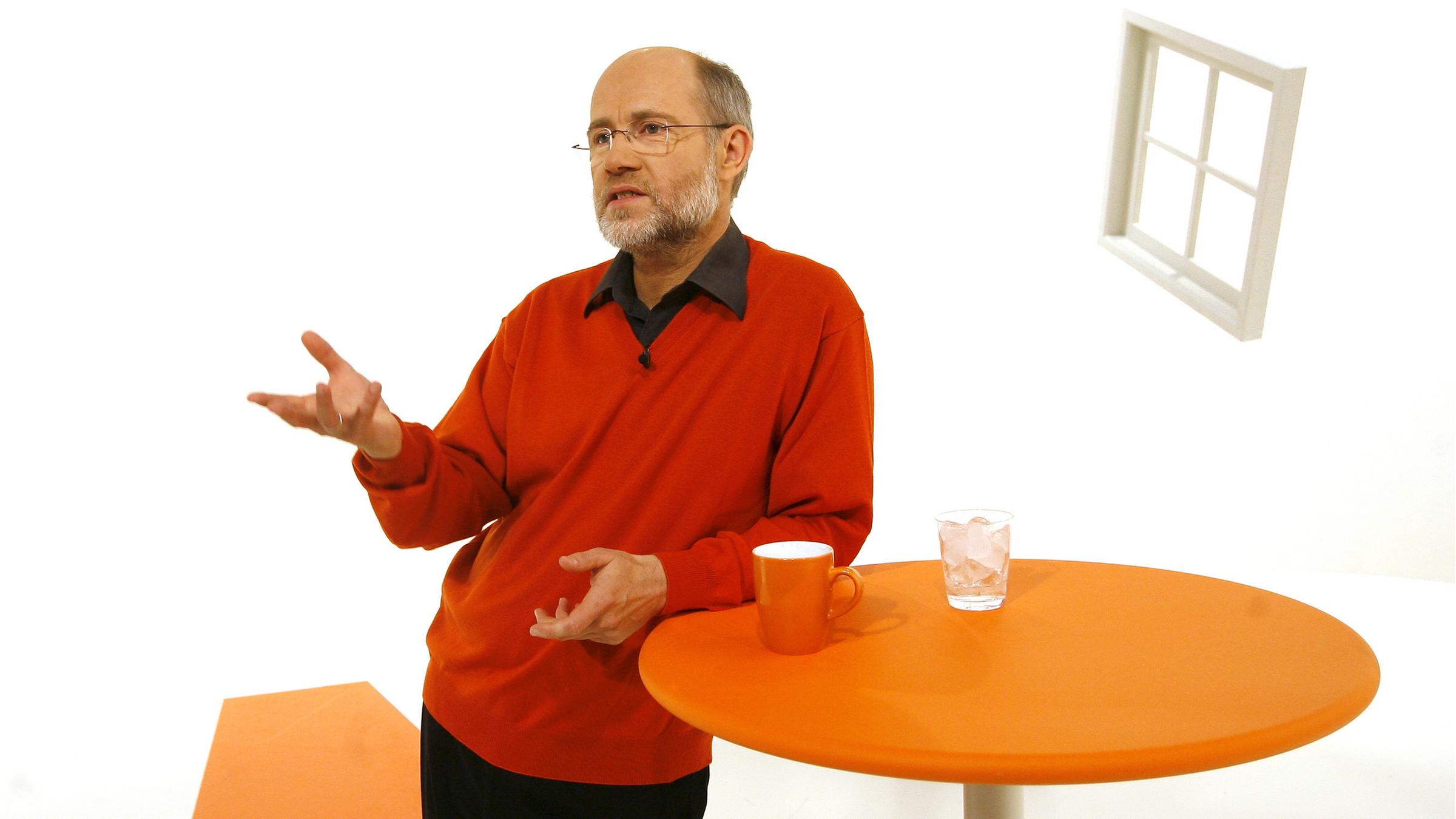 Professor Harald Lesch lehnt an einem orangefarbenen Stehtisch auf dem ein oranger Kaffee-Pot und ein Glas mit Eiswürfeln steht. Auf dem weißen Hintergrund schwebt ein weißes Fenster schräg durch den Raum.