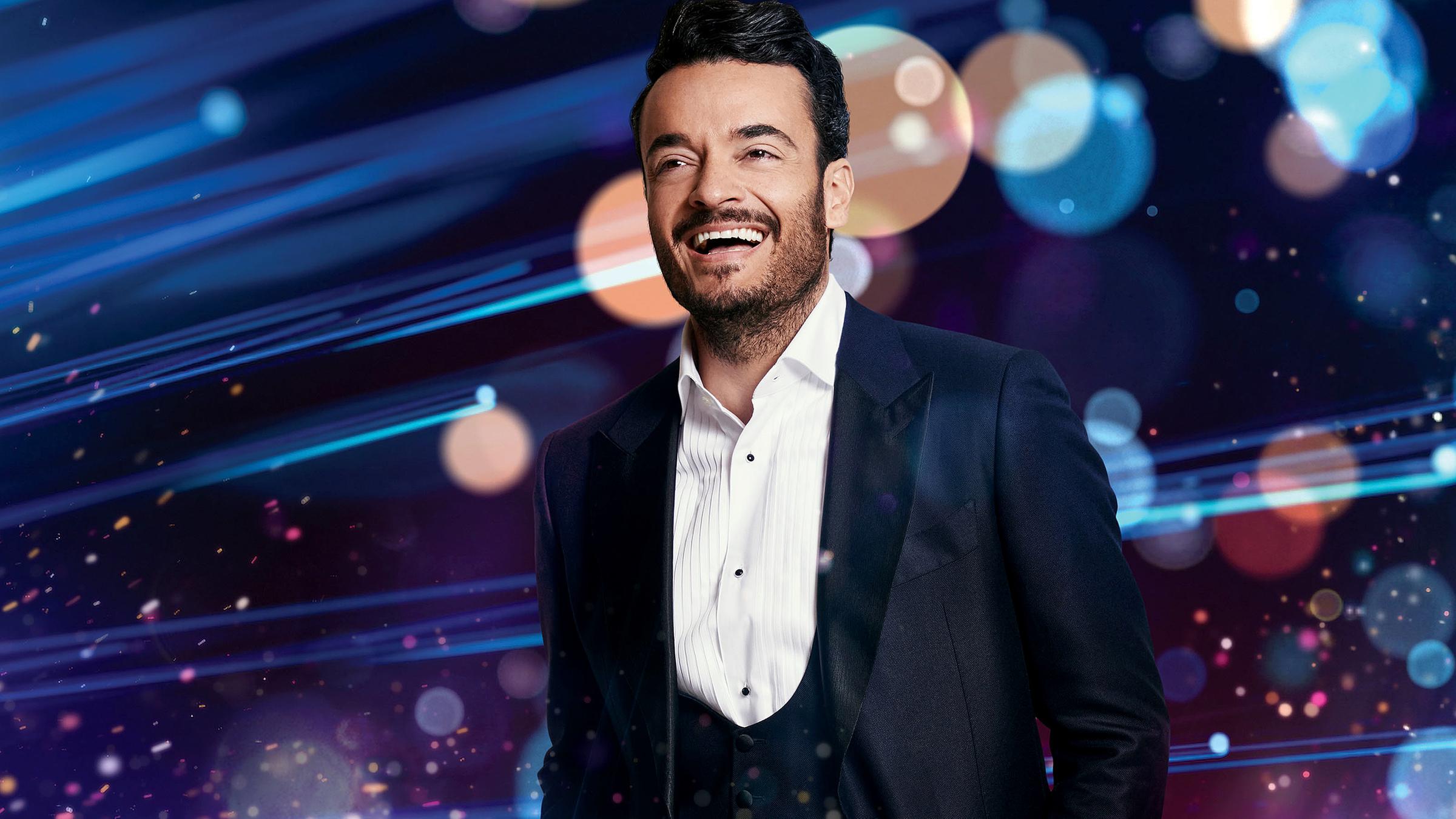 Giovanni Zarrella steht in einem dunklen Anzug vor Leuchteffekten und lächelt.