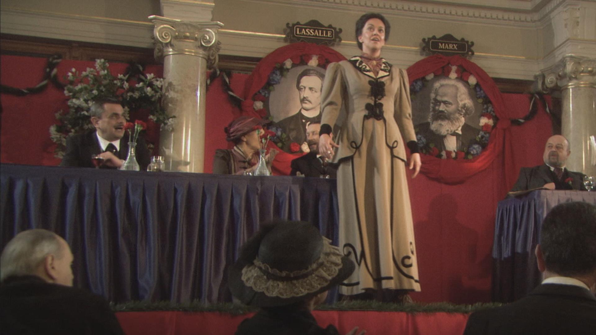 "Die glorreichen 10 - Die glorreichsten Gründer der Geschichte": Spielszene: Rosa Luxemburg steht auf einer Bühne. Vor ihr sitzt das Publikum, hinter ihr stehen zwei Tische, an denen Politiker und Politikerinnen sitzen. An der Wand hängen zwei Bilder von Ferdinand Lassalle und Karl Marx.