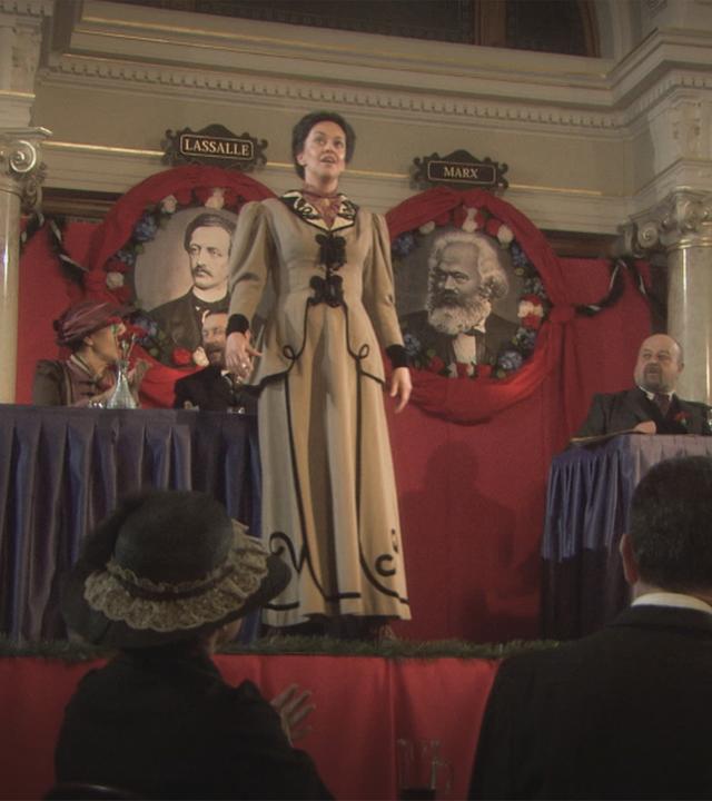 "Die glorreichen 10 - Die glorreichsten Gründer der Geschichte": Spielszene: Rosa Luxemburg steht auf einer Bühne. Vor ihr sitzt das Publikum, hinter ihr stehen zwei Tische, an denen Politiker und Politikerinnen sitzen. An der Wand hängen zwei Bilder von Ferdinand Lassalle und Karl Marx.