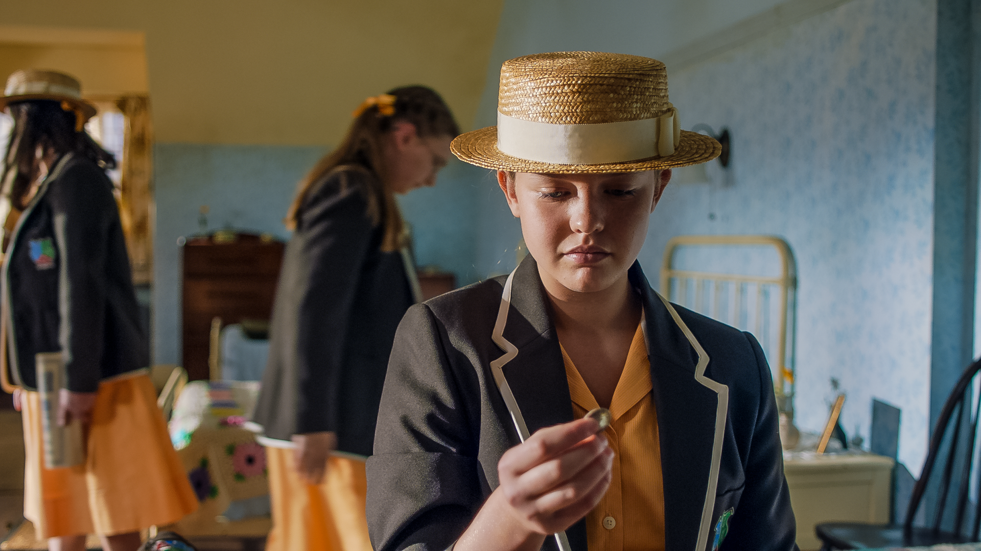 Gwen steht im Schlafsaal und hat ihren Blick von den anderen Mädchen im Raum abgewendet. Heimlich hält sie eine Münze in ihrer Hand und beobachtet diese mit hämischem Blick. Auf ihrem Kopf trägt sie eine Strohhut.