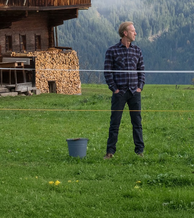 Herbert Sattler (Oliver Stritzel) steht mit den Händen in den Hosentaschen auf der Wiese vor einer Berghütte und blickt nachdenklich zur Seite.