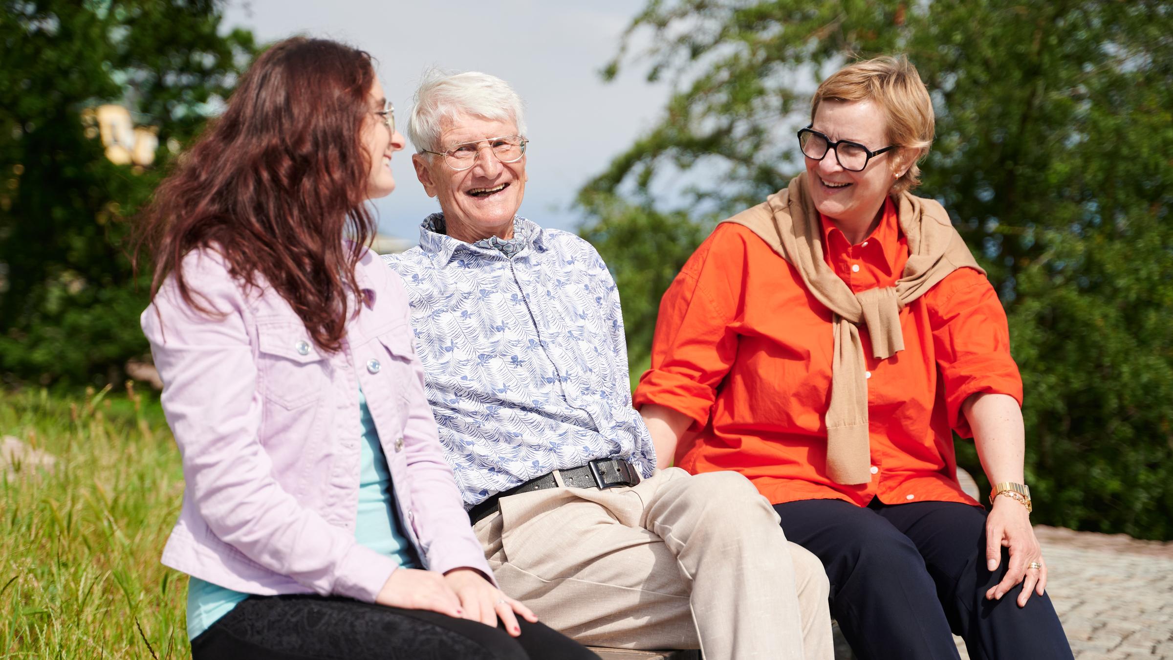 Zwei Frauen und ein Mann sitzen auf einer Bank im Grünen und lächeln sich an.