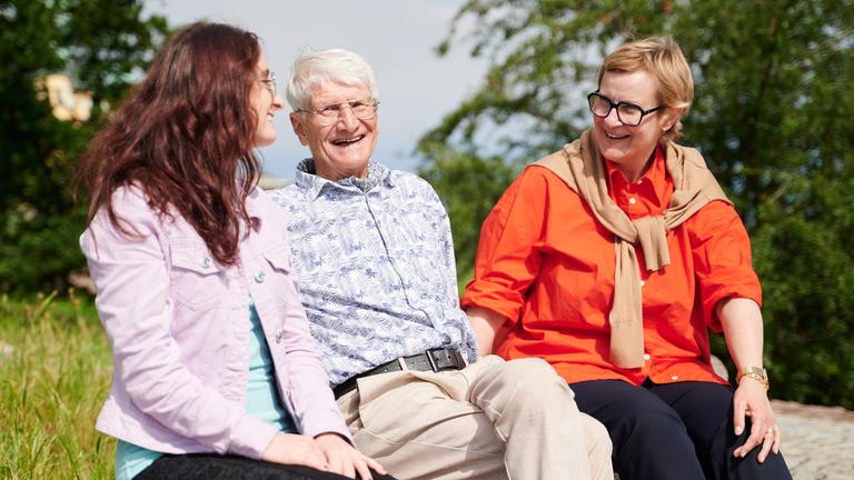 Zwei Frauen und ein Mann sitzen auf einer Bank im Grünen und lächeln sich an.