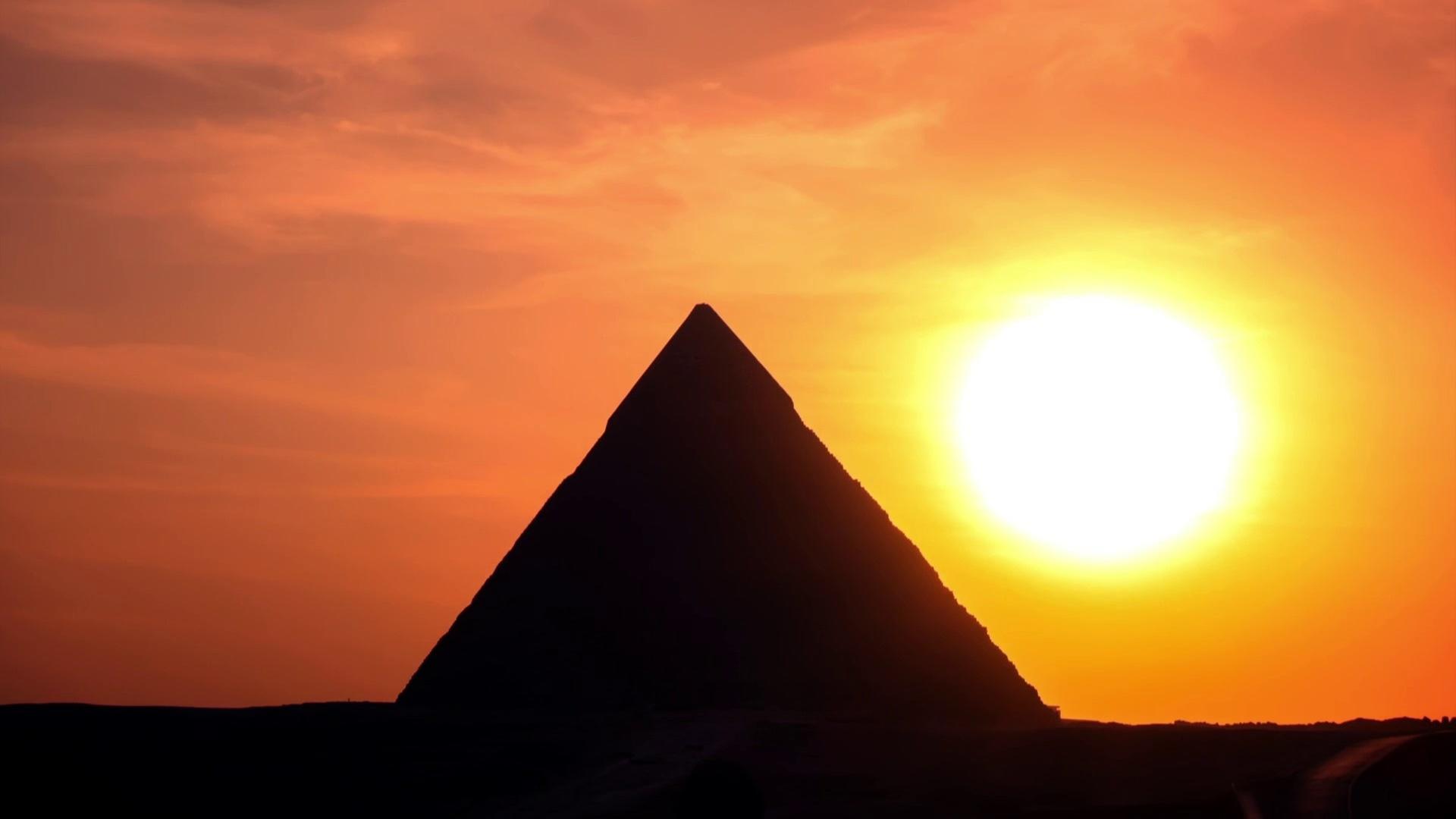 Eine Pyramide im Gegenlicht bei Sonnenuntergang. Die Pyramide wirkt schwarz, die große Sonne rechts daneben leuchtet gelb