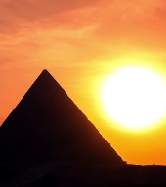 Eine Pyramide im Gegenlicht bei Sonnenuntergang. Die Pyramide wirkt schwarz, die große Sonne rechts daneben leuchtet gelb