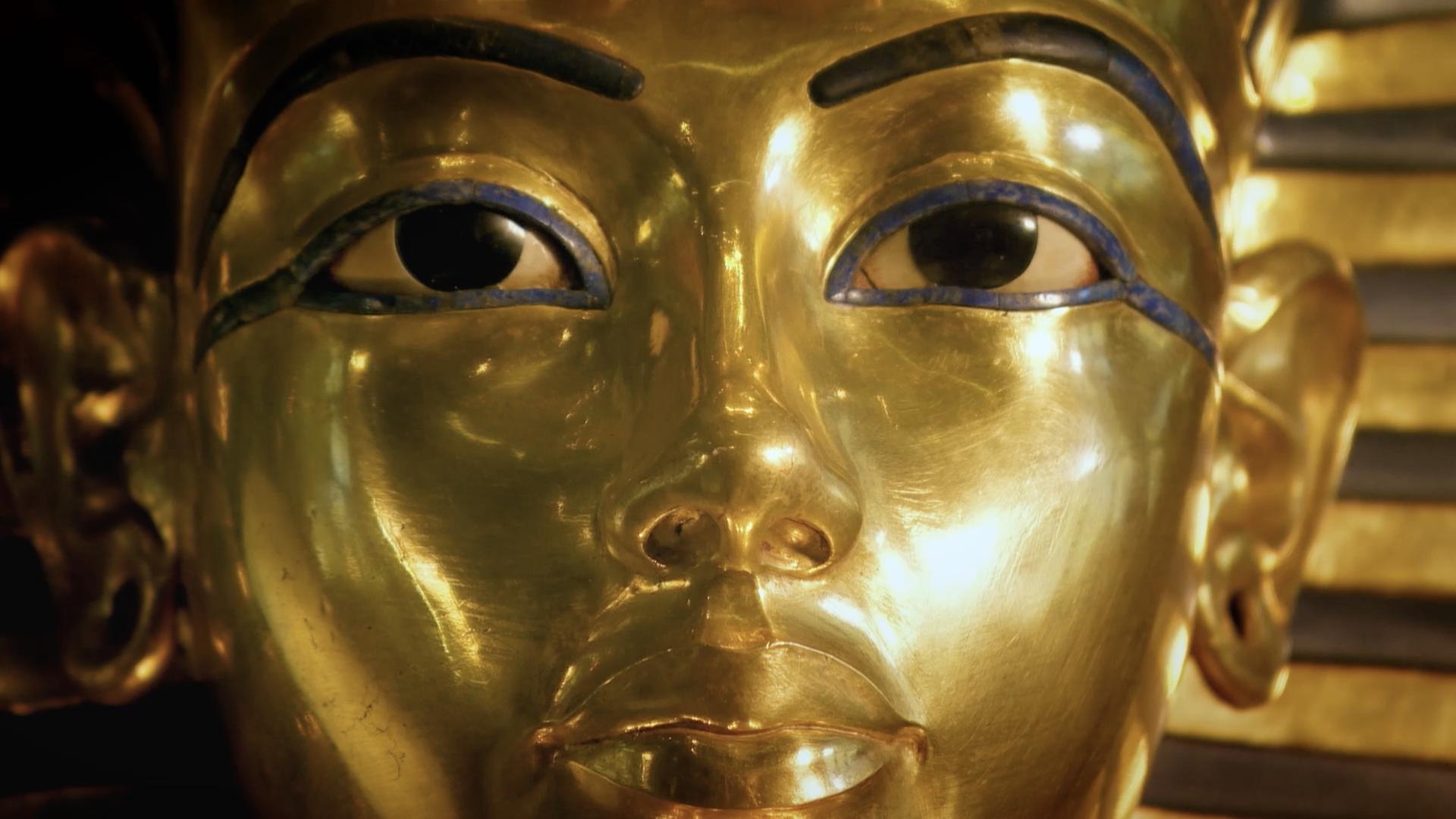 Nahaufnahme. Eine goldene Totenmaske eines Pharaos mit einem Kopfschmuck aus blauen und goldenen Streifen.