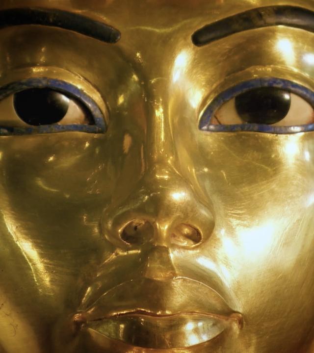 Nahaufnahme. Eine goldene Totenmaske eines Pharaos mit einem Kopfschmuck aus blauen und goldenen Streifen.