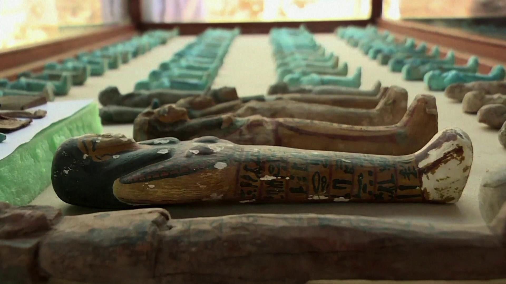 Viele kleine Schabtis, kleine Mumien-Figuren, liegen in mehreren Reihen nebeneinander auf einer Fläche. Ein Teil ist mit blauer Fayence glasiert.