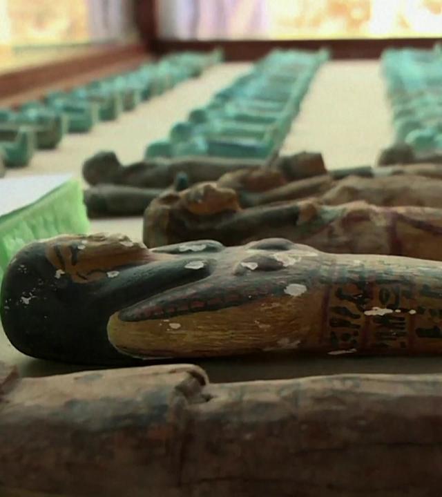 Viele kleine Schabtis, kleine Mumien-Figuren, liegen in mehreren Reihen nebeneinander auf einer Fläche. Ein Teil ist mit blauer Fayence glasiert.