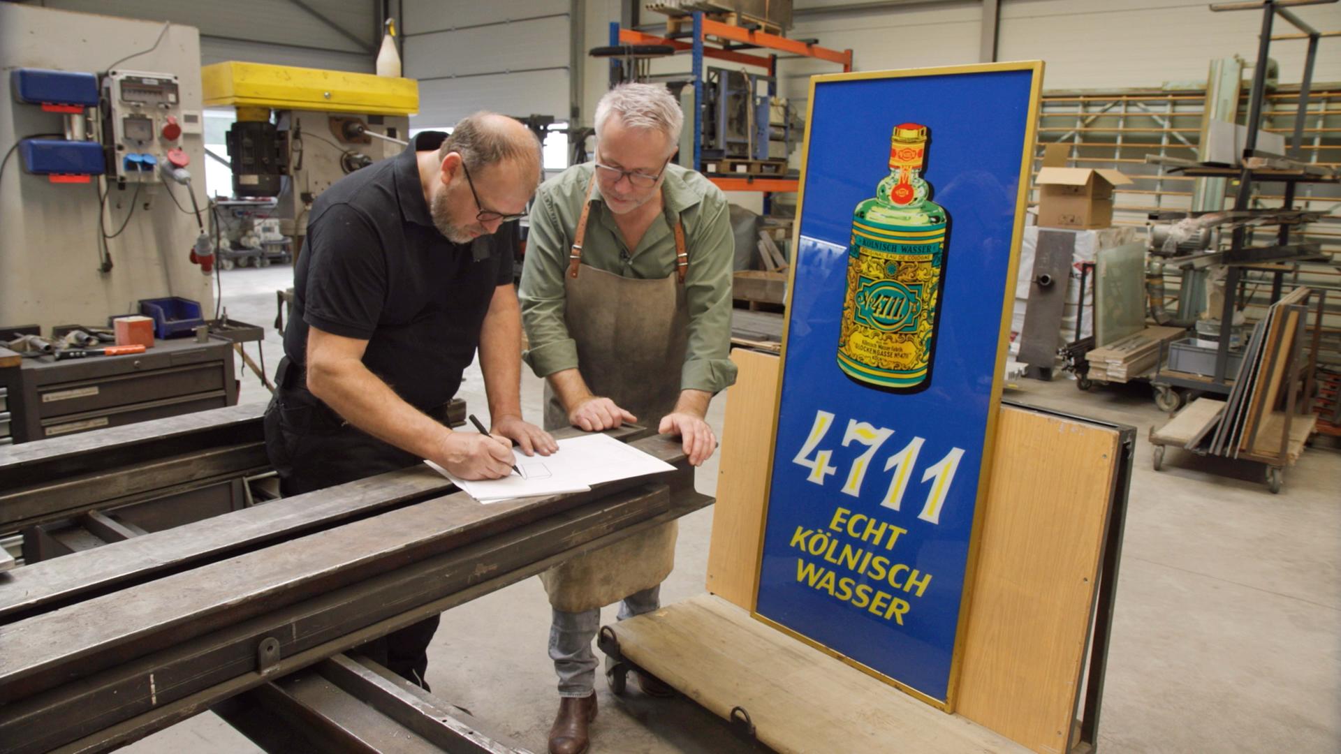 Andreas Köpp und Jan Cizek entwerfen eine Skizze. Daneben steht ein altes Werbeschild aus Glas für Kölnisch Wasser.