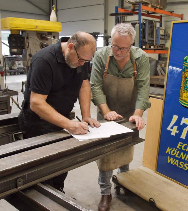 Andreas Köpp und Jan Cizek entwerfen eine Skizze. Daneben steht ein altes Werbeschild aus Glas für Kölnisch Wasser.