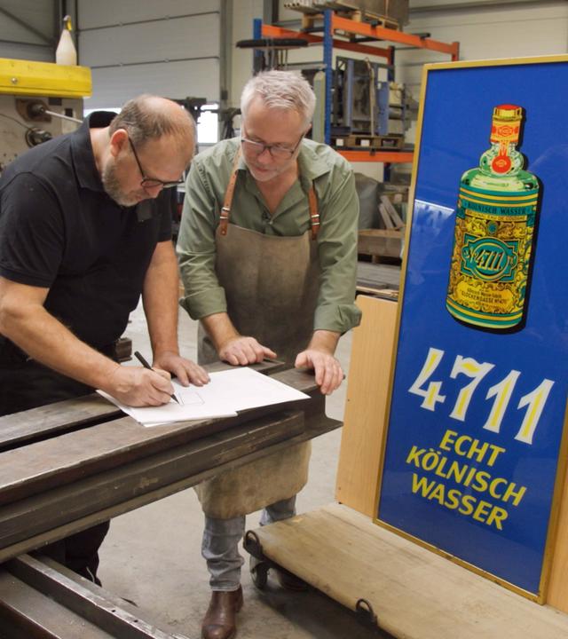 Andreas Köpp und Jan Cizek entwerfen eine Skizze. Daneben steht ein altes Werbeschild aus Glas für Kölnisch Wasser.