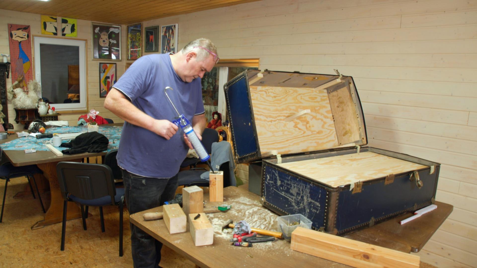 Waldi repariert mit einer Klebepistole einen alten Überseekoffer.