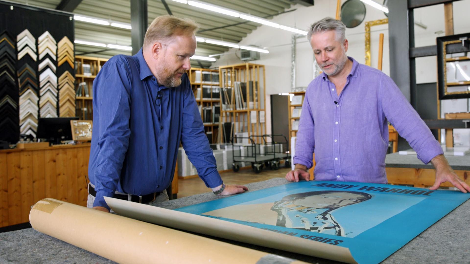 Händler Jan Čížek und ein Experte für Rahmen schauen sich ein auf dem Tisch ausgebreitetes Andy Warhol Poster aus der Ausstellung "The American Indian Series" an.