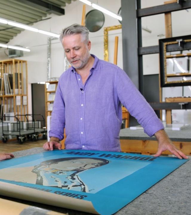 Händler Jan Čížek und ein Experte für Rahmen schauen sich ein auf dem Tisch ausgebreitetes Andy Warhol Poster aus der Ausstellung "The American Indian Series" an.