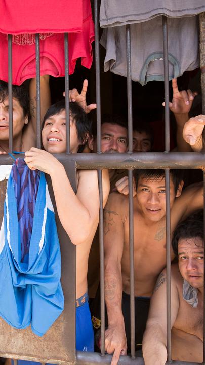 Fünf Männer mit nacktem Oberkörper zwängen sich hinter einer vergitterten Türe und schauen nach draußen.