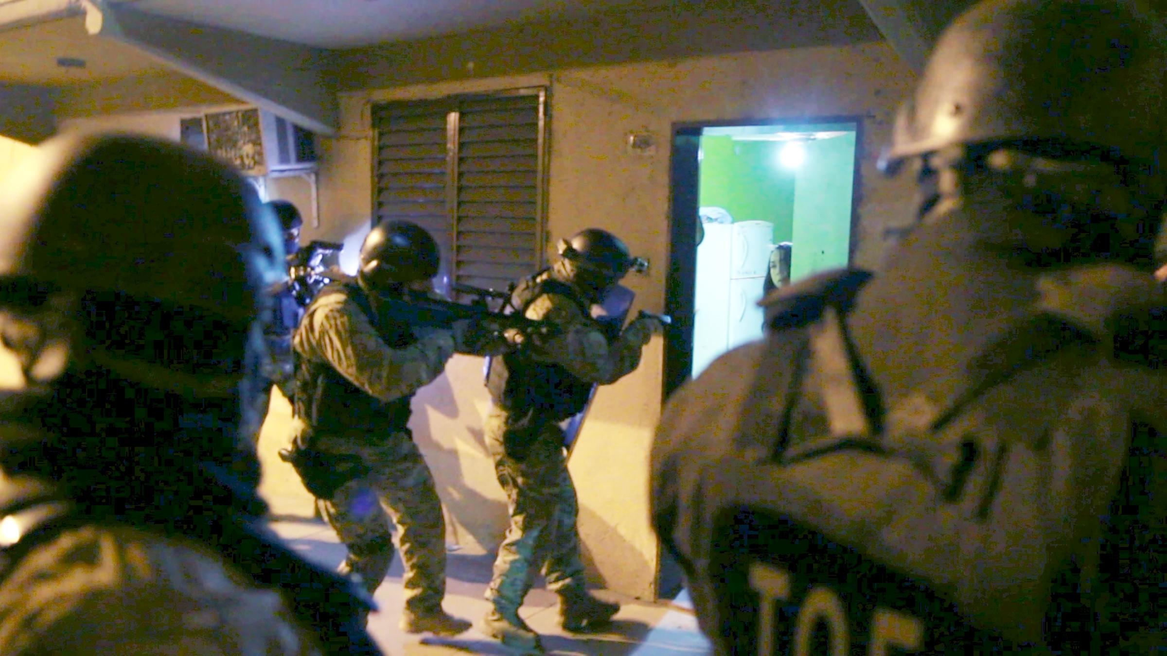 Polizisten mit Helm und Kampfausrüstung stehen vor einer geöffneten Türe. Sie halten Schusswaffen im Anschlag.