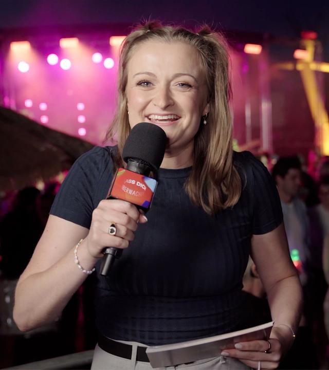 Anna Neudecker moderiert "Die halbe Stunde danach". Im Hintergrund sind viele Menschen bei einer Party unter freiem Himmel zu sehen.