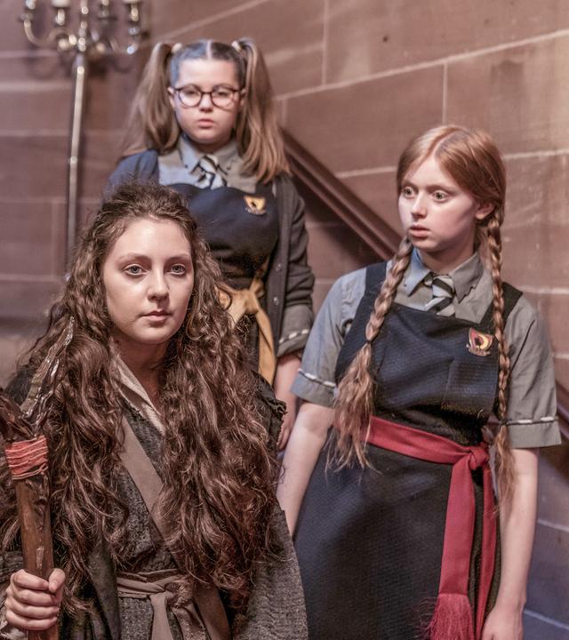 In einem alten gemauerten Treppenhaus steht Mildred (rechts) mit ihren beiden Freundinnen Edith und Maude um "Gertrude, die Große" herum. Gertrude hat einen Magierstab aus dunklem Holz in ihrer rechten Hand.