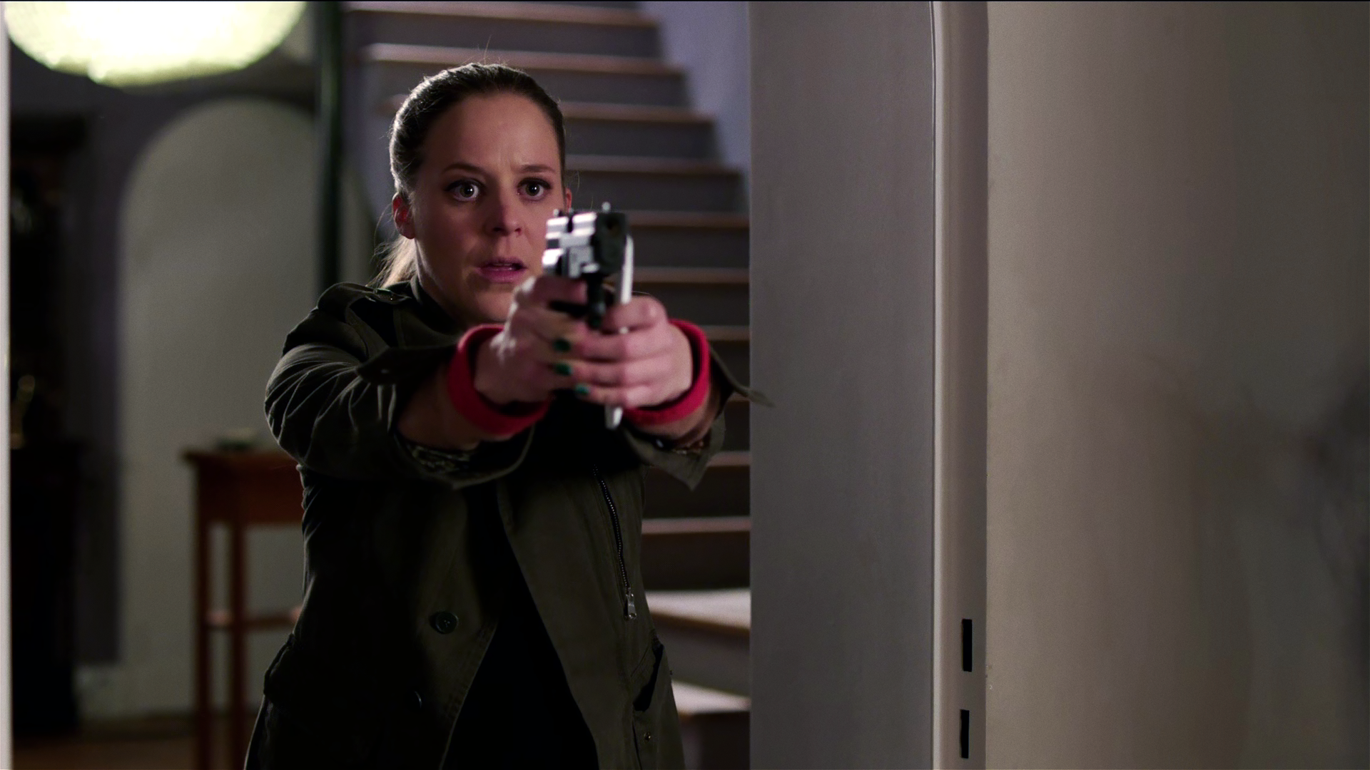 In einem Hausflur steht Angelika Flierl (Bernadette Heerwagen). Sie sieht erschrocken aus und zielt mit ihrer Waffe. 