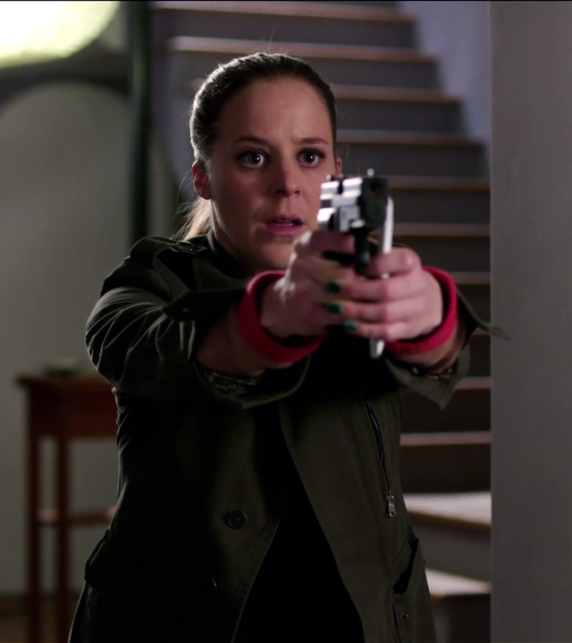 In einem Hausflur steht Angelika Flierl (Bernadette Heerwagen). Sie sieht erschrocken aus und zielt mit ihrer Waffe. 