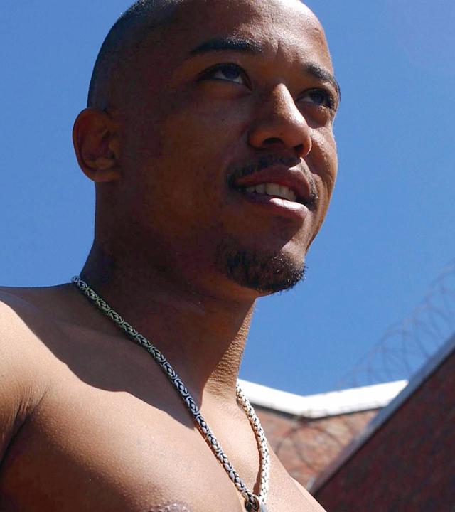 Nahaufnahme eines jungen Mannes mit nacktem Oberkörper. Im Hintergrund Gefängnismauern mit Stacheldraht.