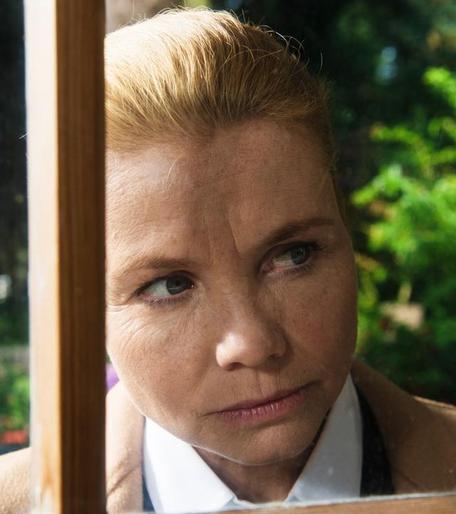 "Ella Schön - Die Inselbegabung": Ella (Annette Frier) schaut durch eine Fensterscheibe ins Innere eines Hauses. Hinter ihr sind verschwommene Ausschnitte eines Vorgartens erkennbar.