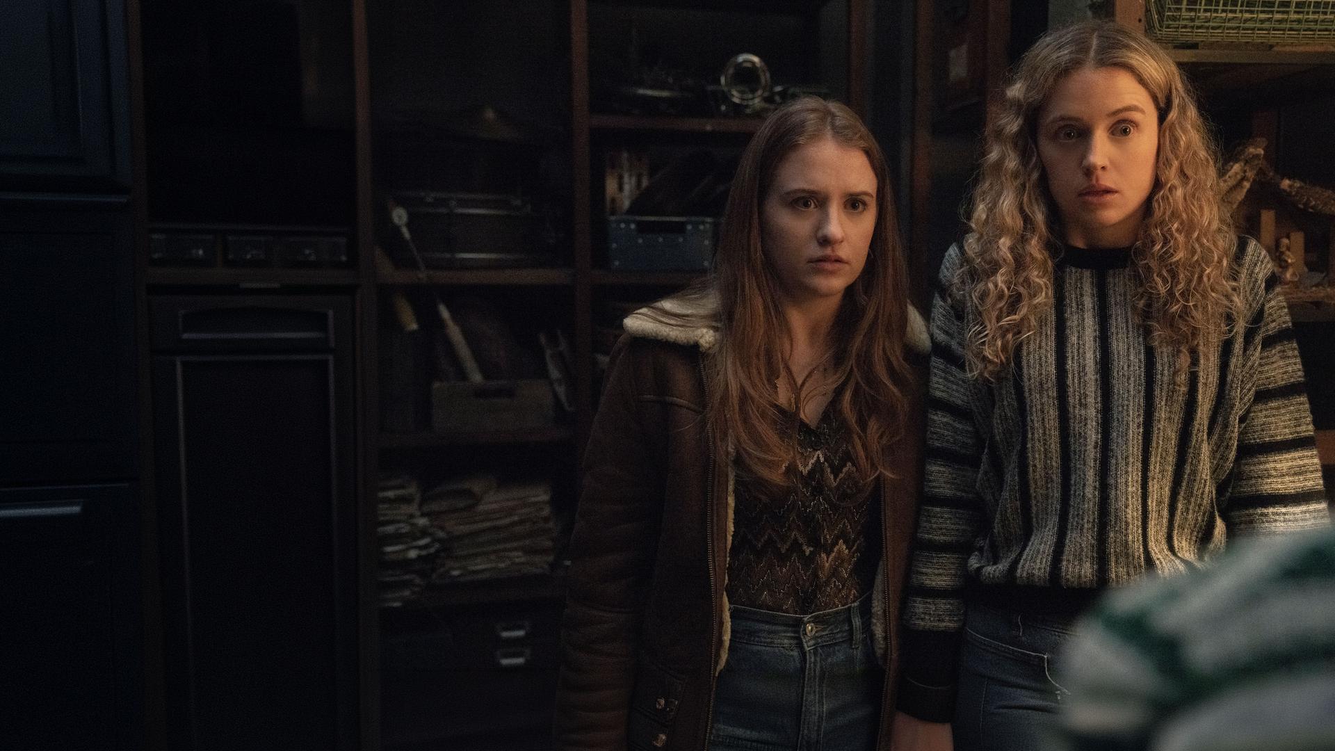 "Extraordinary - Die Jen-Show": Jen (Máiréad Tyers) und Carrie (Sofia Oxenham) stehen in einem Raum mit hohen Regalen und blicken schockiert geradeaus.
